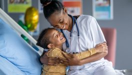 A sweet little pediatric patient, of African decent, sits on the edge of his hospital bed as he gives his nurse a warm hug.  He is dressed comfortably in pajamas and looking up at his nurse with a big smile.